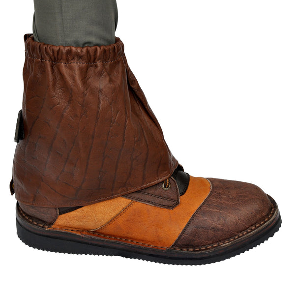 Side view of a Buffalo Game Skin Gaiter, color Brown. The gaiter has an elastic band and brass snap at the top, a Velcro secured leather tab strap with a Tag Safari logo in the middle, a brass snap at the bottom, and a reinforced stitched seam that runs vertically from top to bottom. Genuine game skin leather.