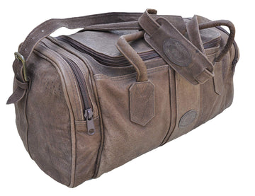 Front and left side of a Tag Safari Warthog Leather Duffle in Brown. Shows a full zippered top opening, zippered side pouches, an adjustable padded shoulder strap, cushioned handles, and an embossed Tag logo leather patch.